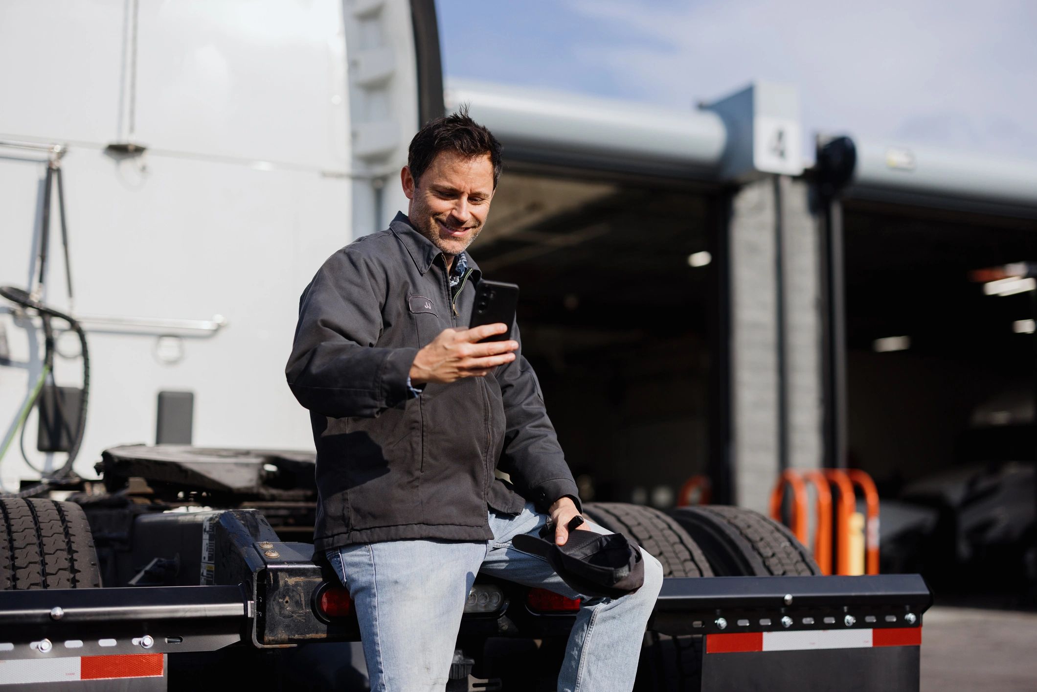 Man in jacket sitting on truck chassis using smartphone.