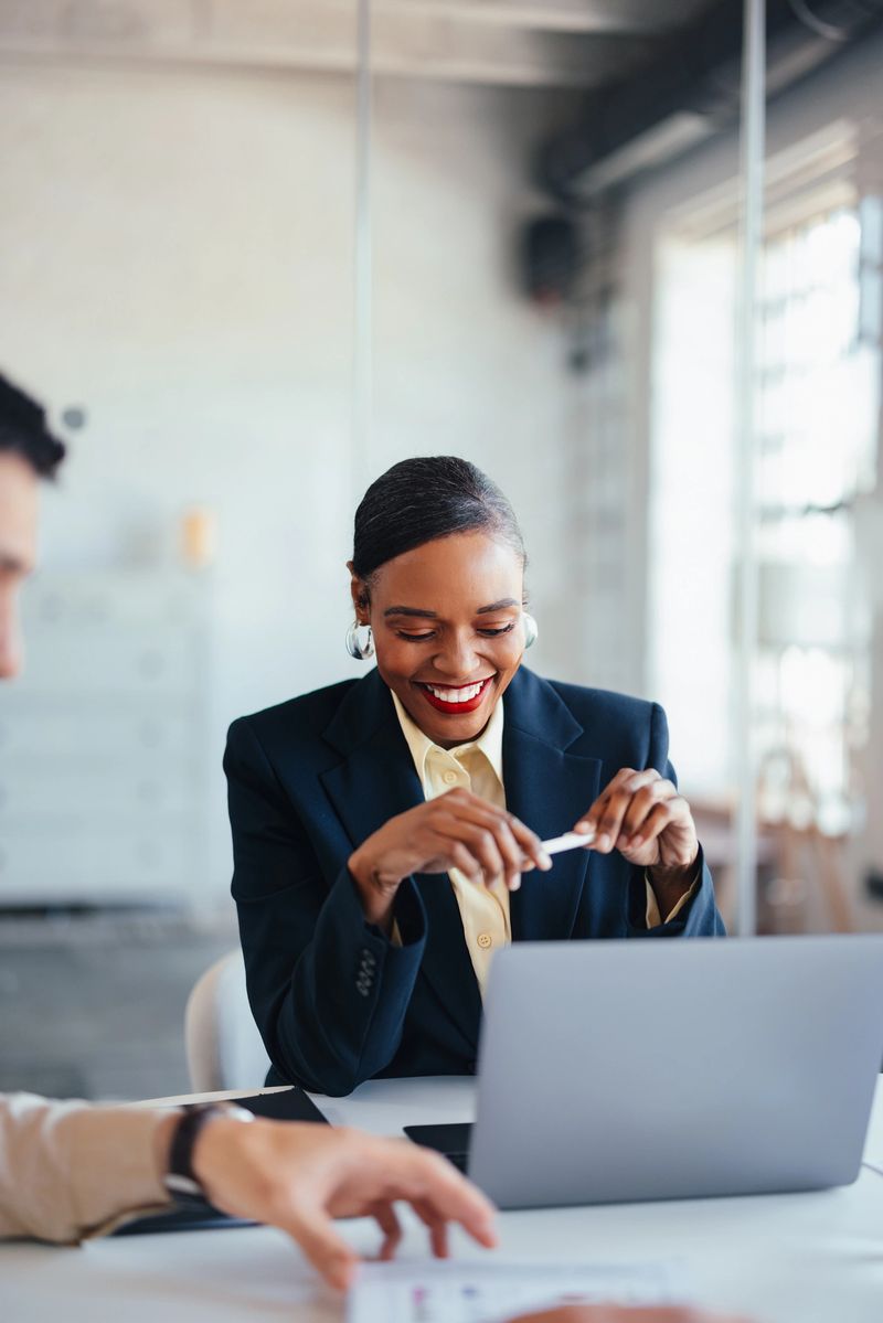 A professional woman in a navy blazer smiles as she prepares documents at a modern office, using a laptop. The scene conveys collaboration, focus, and positive teamwork in a corporate setting.