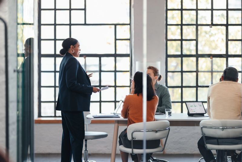 A diverse team gathers around a conference table as a confident speaker presents notes in a sunlit office. Laptops and charts indicate collaboration, strategy planning, and a dynamic business environment.