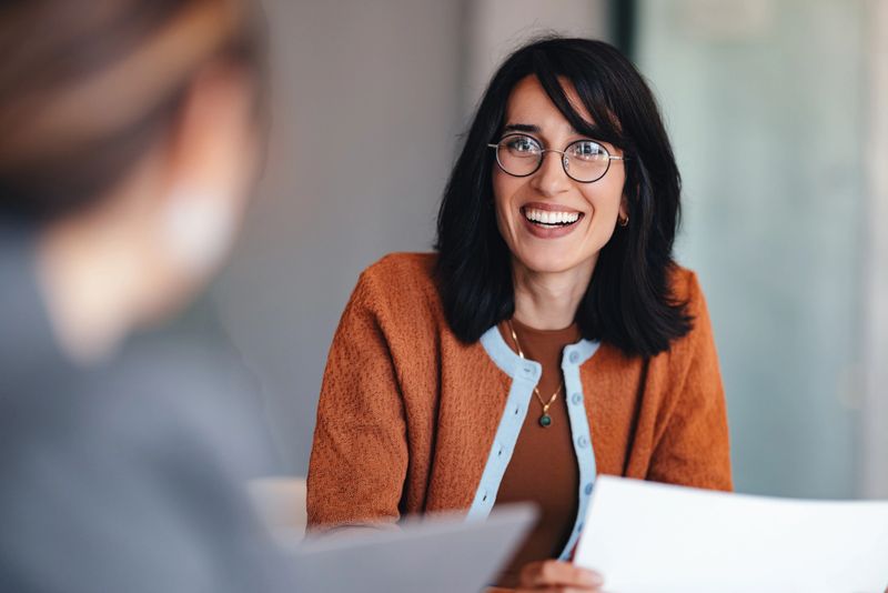 A friendly professional woman with glasses smiles while sharing documents during a casual office meeting. The scene conveys collaboration, communication, and positive teamwork in a modern workspace.
