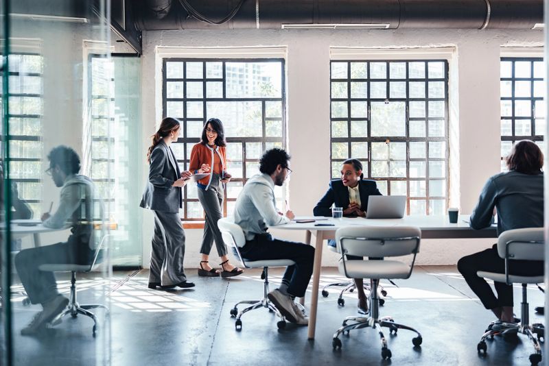 A diverse group of professionals gathers around a table in an airy, glass-walled office.