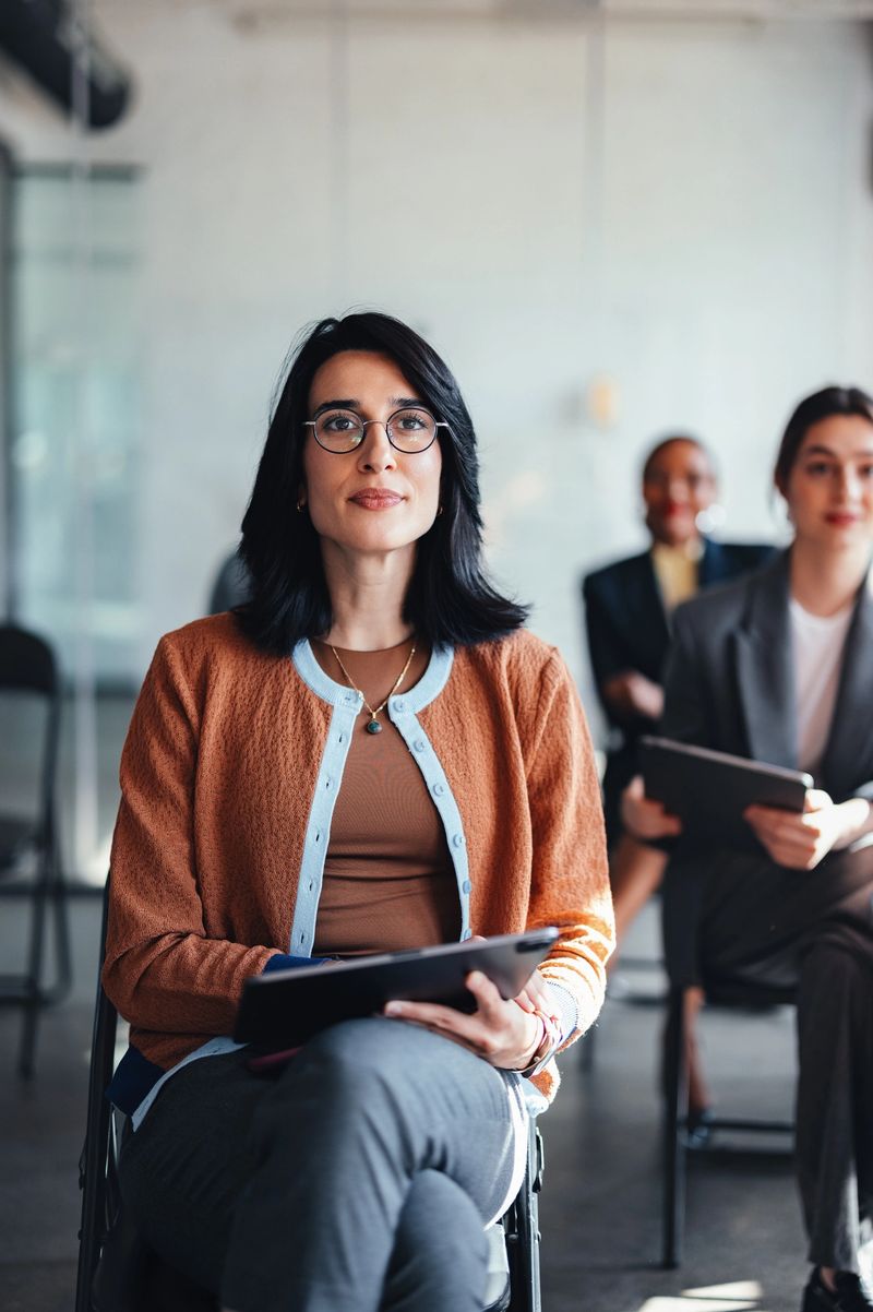 A poised woman wearing glasses and an orange cardigan sits in a contemporary office, with colleagues in the background. The scene communicates professionalism, teamwork, and a corporate atmosphere.