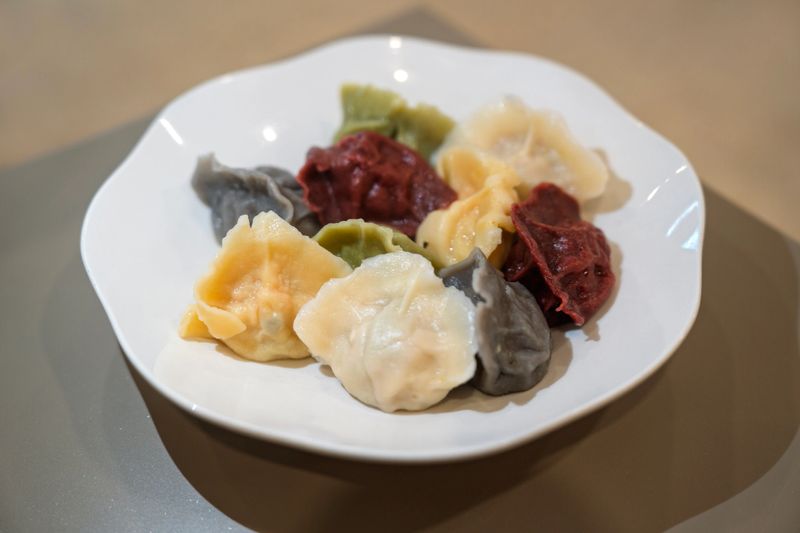 A close-up view of assorted multicolored dumplings served on a white plate. The vibrant dumplings feature various hues such as red, green, grey, and white, showcasing a variety of flavors and ingredients. Captured indoors, this image highlights Asian cuisine and a ready-to-eat meal.