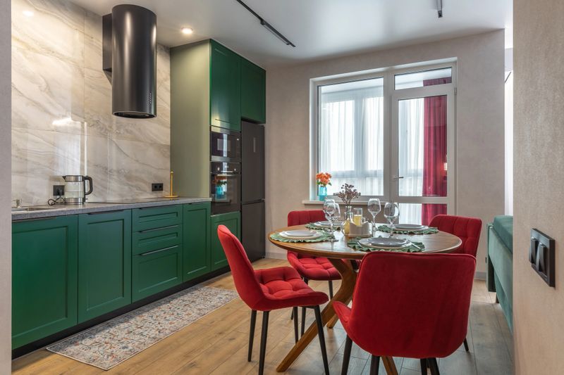 Bright modern kitchen interior with green cabinets, red dining chairs, wooden table, and elegant table setting in cozy apartment.