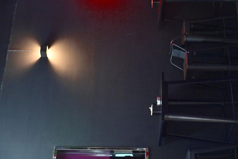 Authentic interior scene of dark wall with upward and downward casting sconce creating conical light patterns, high table with dish and white object, two bar stools, and colorful ambient reflections.