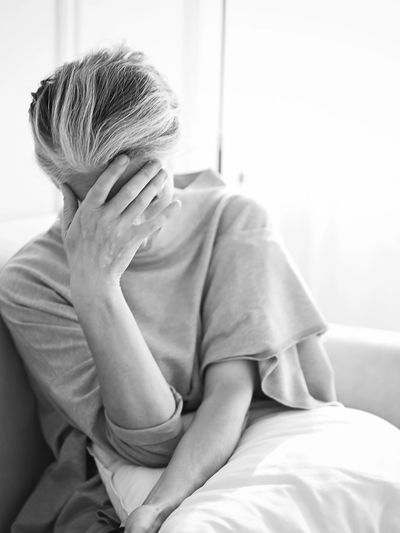 An elderly woman sitting on a couch with her face in her hand, appearing distressed.