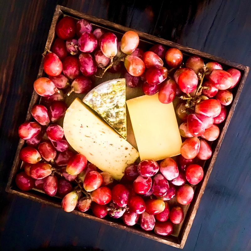Top-down artistic composition of fresh red grapes and artisan cheeses arranged on a wooden board. Warm rustic styling evokes slow living, hospitality, and Mediterranean farmhouse dining.