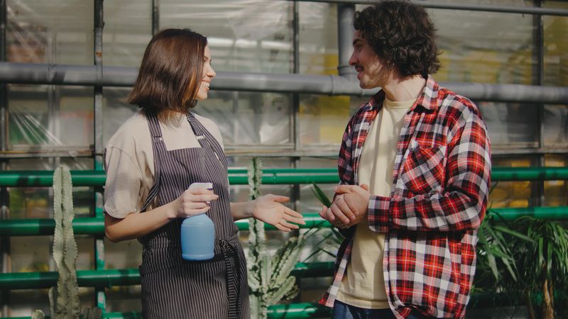 Two individuals are talking happily in a greenhouse, surrounded by green plants. One holds a spray bottle, likely discussing plant maintenance techniques.