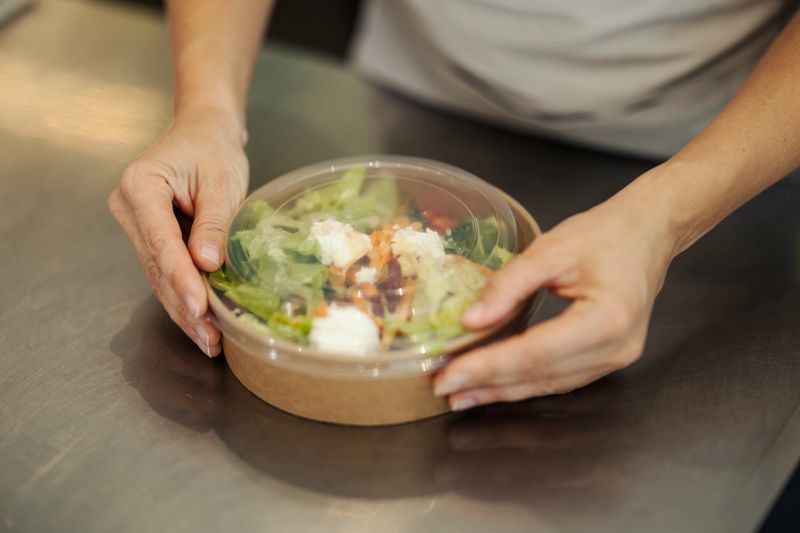 Hands placing lid on a fresh, healthy salad in a sustainable take-out container on a metal surface, conveying healthy eating and food service