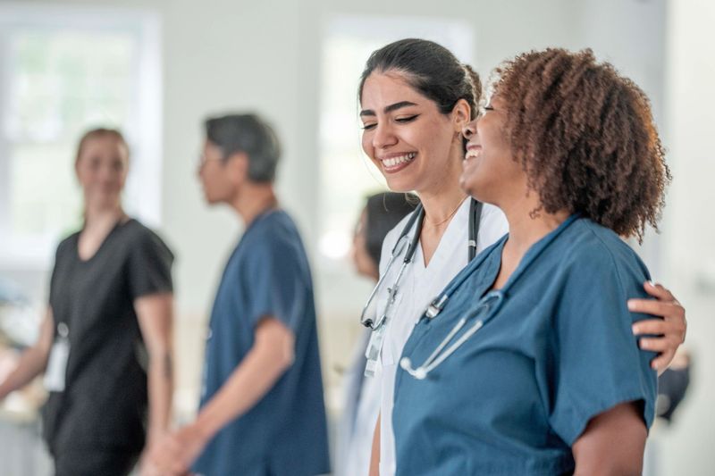A diverse group of healthcare professionals in a bright hospital setting shares a lighthearted moment, demonstrating teamwork, camaraderie, and support among doctors and nurses across different backgrounds.