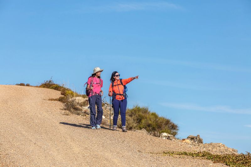 Two female hikers pause along a gravel path to discuss their route, with one pointing forward. The bright blue sky and natural surroundings create an uplifting, adventurous atmosphere.