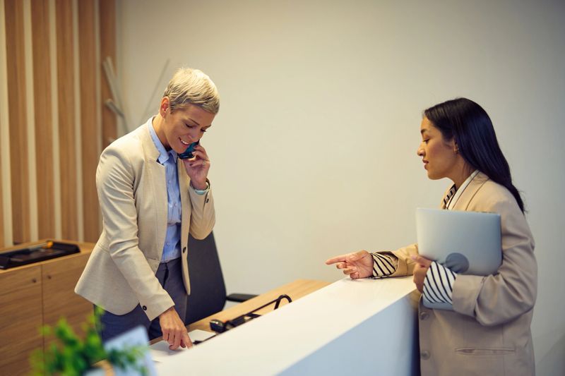 Professional receptionist talking on the phone and pointing at documents while assisting a businesswoman carrying a laptop at the office reception area