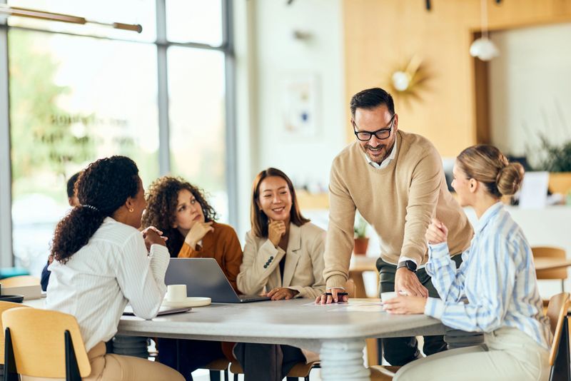 A diverse group collaborates around a round table while a confident leader guides discussion.