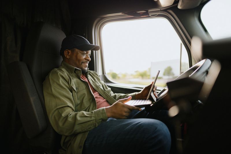 Smiling mature African American truck driver working on a laptop and digital tablet while sitting comfortably inside the truck cabin