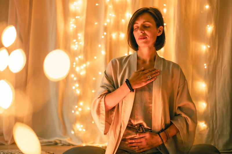 Peaceful woman meditates with hands on heart and abdomen. Gentle golden lights create harmony and calm, symbolizing self-connection, feminine energy, mindfulness, and inner healing.