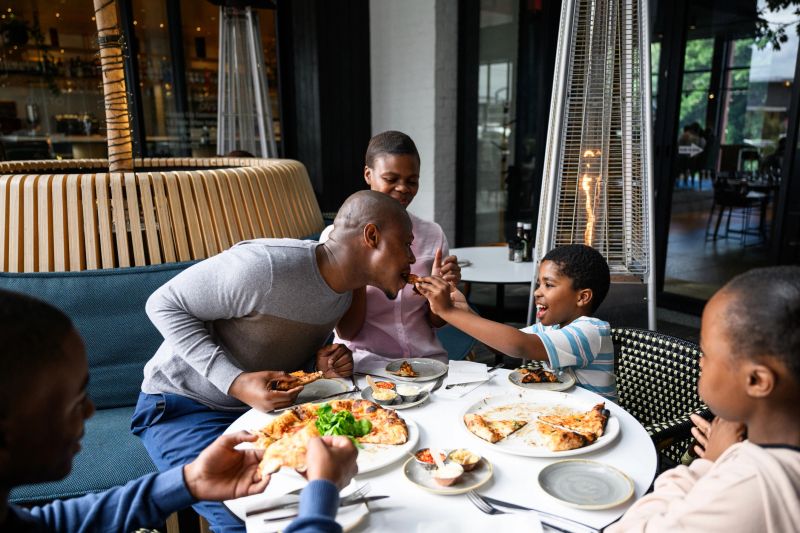 Father leaning towards his son to bite a pizza slice while dining together around a restaurant table, expressing bonding, enjoyment and quality family time during a relaxed meal.