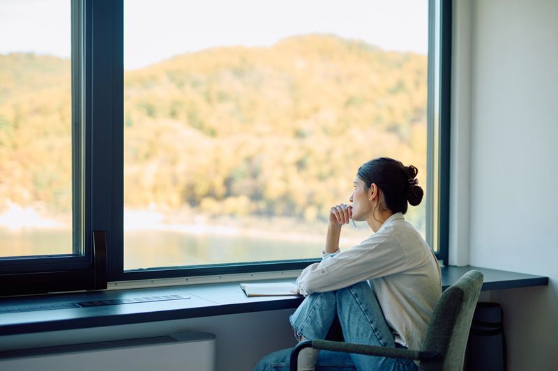Young woman sitting by a large window, looking out at the autumn landscape and lake, thinking or contemplating