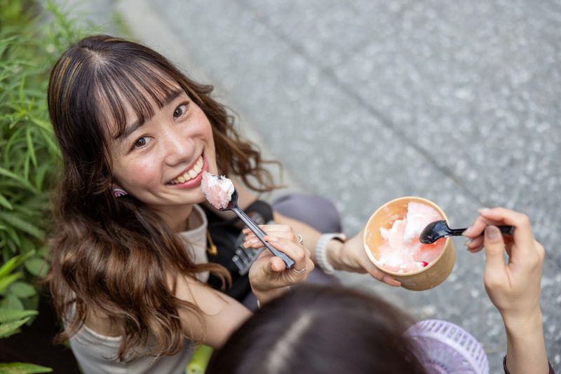 Young female friends sitting on street and eating ice cream at Matsuri, traditional Japanese festival - looking at camera