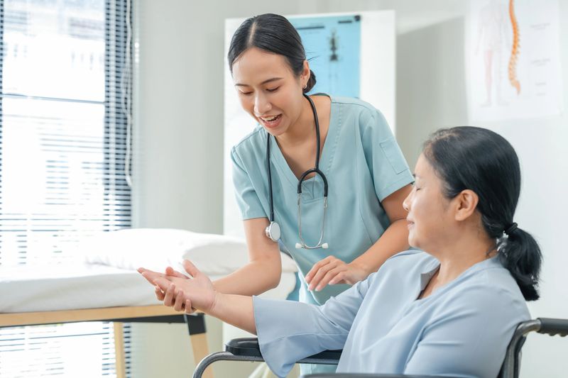 Asian nurse gently holds the hand of a senior patient in a wheelchair, offering emotional comfort and compassionate care. The warm connection between caregiver and elderly person reflects trust