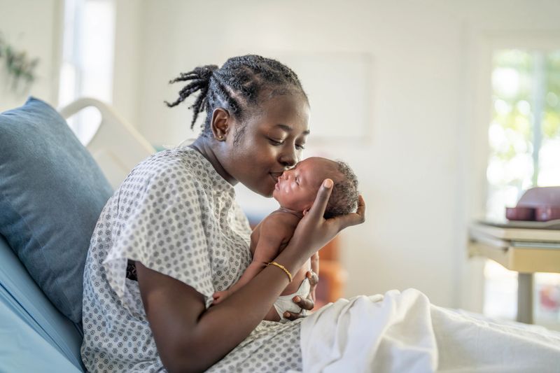A Black mother in a hospital bed cradles and kisses her newborn, capturing a intimate moment of love, bonding, and family care in a quiet, hopeful hospital setting.