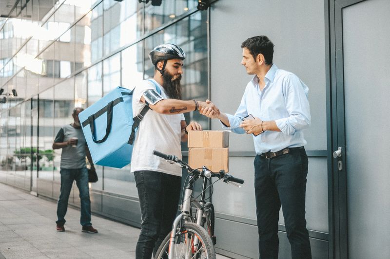 A bearded delivery man on a bicycle, wearing a helmet and carrying a blue bag, hands a package to a businessman in a professional setting with glass buildings in the background.