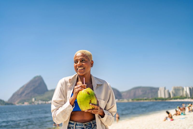 Smiling woman enjoying a fresh coconut drink on a sunny beach with ocean and mountains in the background