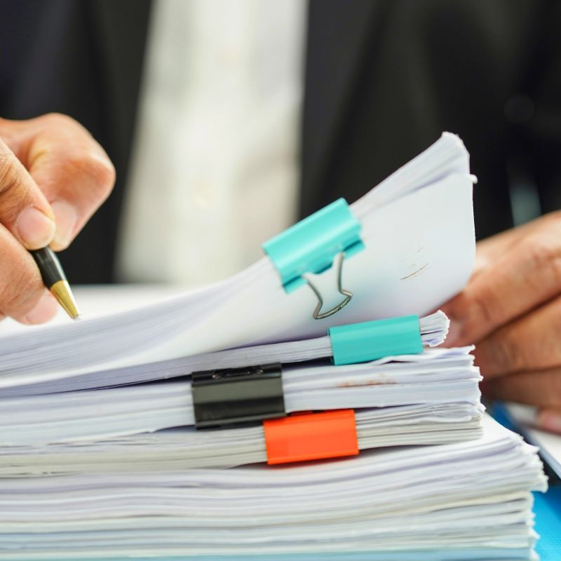 A close-up shot shows a person's hands holding a pen and reviewing a stack of papers secured with colorful clips.