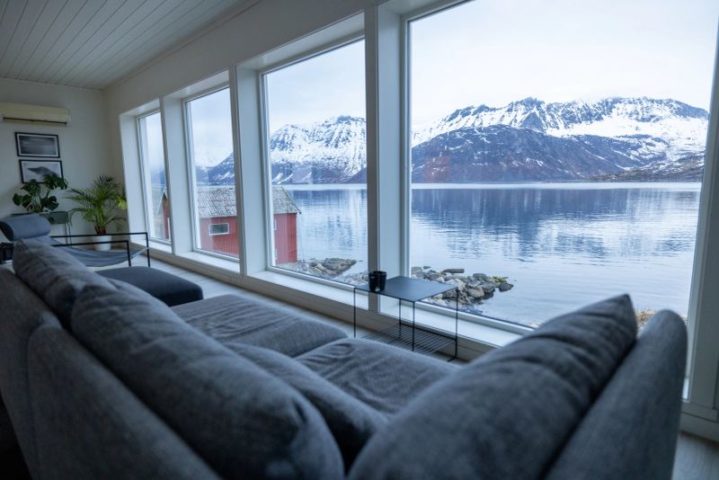 Modern living room with large windows overlooking a calm fjord and snow-capped mountains. Comfortable gray sofa and indoor plants create a peaceful, scenic interior space. Relaxing home with nature view.