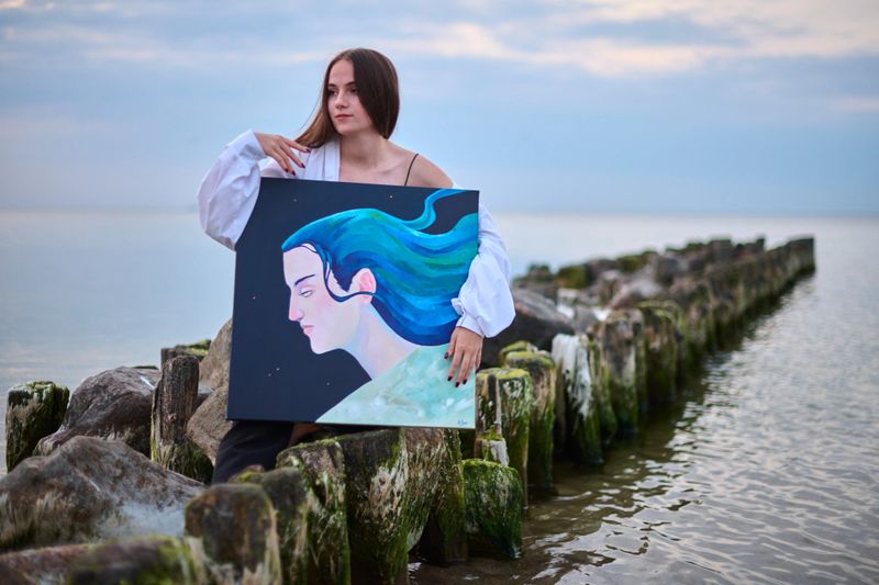 Young woman stands on rocky pier holding portrait she painted, set against calm sea. Painting features serene face with flowing blue hair, reflecting tranquility and creativity
