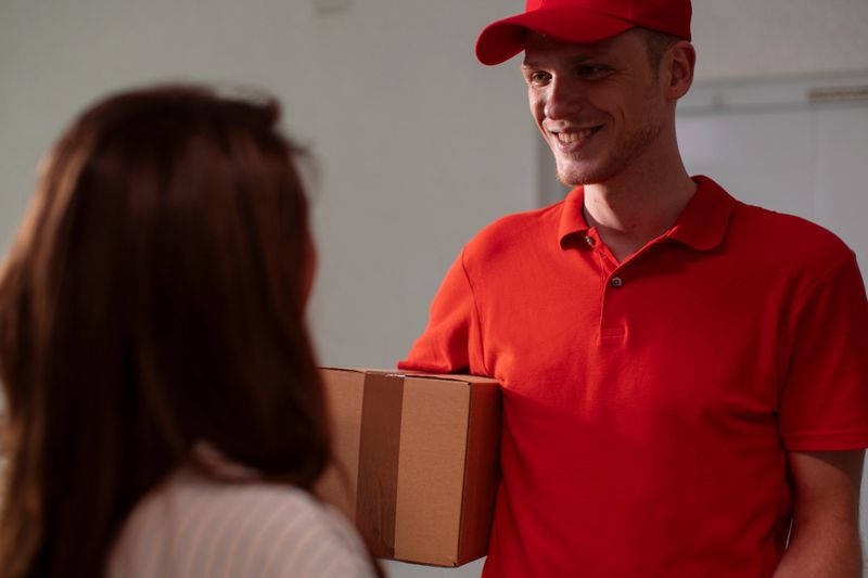 A delivery worker in a bright red polo and cap hands a cardboard package to a customer, conveying friendly service and everyday home-delivery interaction