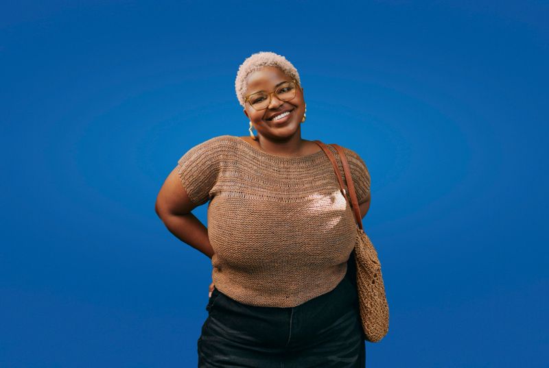 Portrait of a young Black woman with short blonde hair smiling confidently outdoors against a bright solid blue background. She wears glasses, casual brown knit top, black pants, and carries a shoulder bag