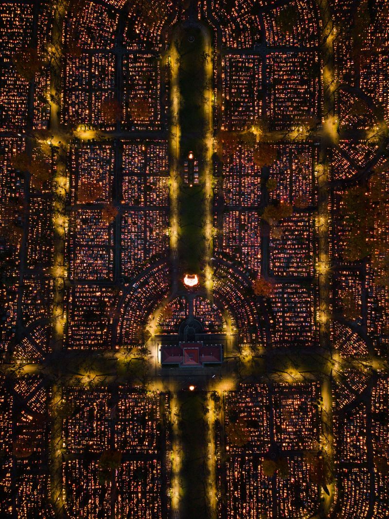 Aerial view of the Central Cemetery in Gliwice, Poland, during All Saints’ Day. Thousands of candles illuminate the graves as families gather to honor their loved ones. The warm lights create a peaceful atmosphere contrasting with the evening cityscape.