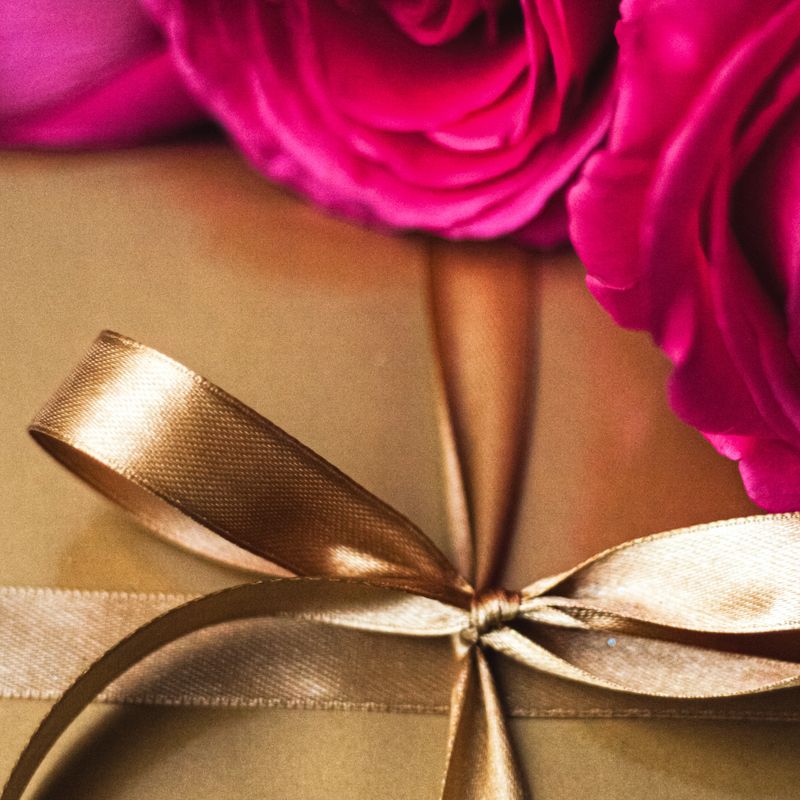 Present box adorned in shimmering golden paper, accented with a luxurious satin ribbon bow and lively pink roses, symbolizing memorable moments