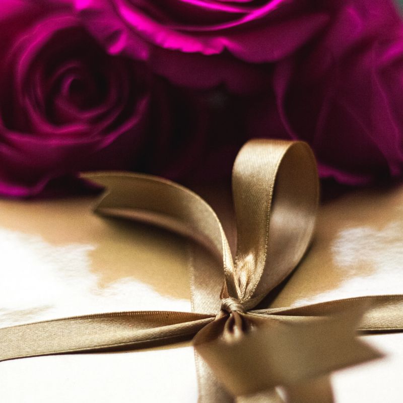 Gift box wrapped with gold ribbon and bow, accompanied by beautiful magenta roses on a dark background