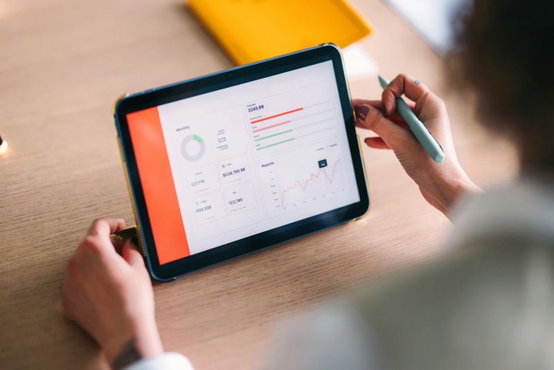 A tablet displays a business dashboard with charts and metrics while a person uses a stylus on a wooden desk, suggesting data analysis, productivity, and digital work in a modern office setting.