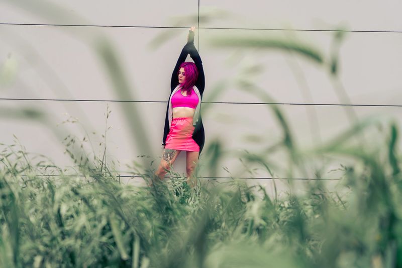 Young woman with pink hair stretching against a modern textured wall, standing among wild urban grasses in bold streetwear style