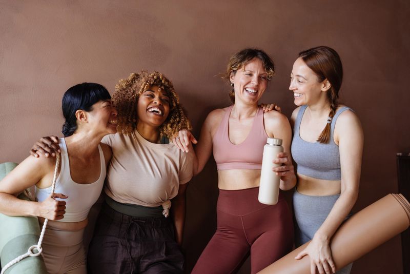 A group of four women in athletic wear share a joyful moment after a workout, showcasing friendship, fitness, and teamwork in a warm studio setting.