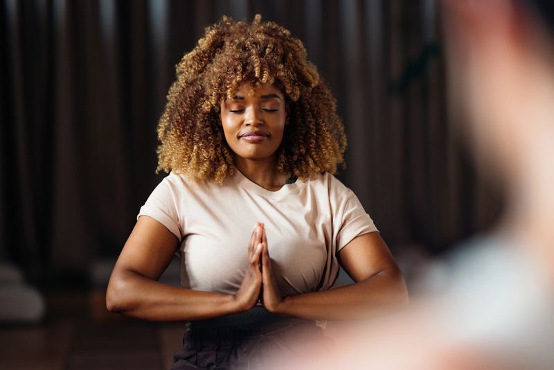 A focused woman sits in a calm yoga class, eyes closed, hands together in a prayer pose, creating a serene moment of mindfulness and balance in a softly lit studio.