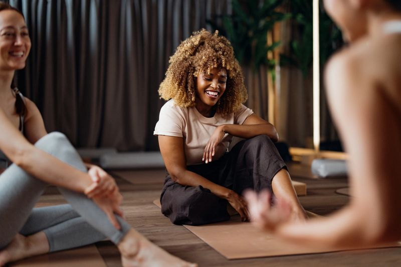 Smiling participants share a relaxed yoga class in a warm studio. A diverse group sits on mats, creating a calm, friendly, and supportive atmosphere for mindful movement and connection.