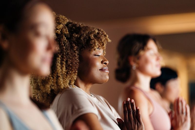 Diverse group of women in a calm, meditative moment, standing with hands in prayer. Warm lighting, serene expressions, focused on inner peace and mindfulness during a yoga class.