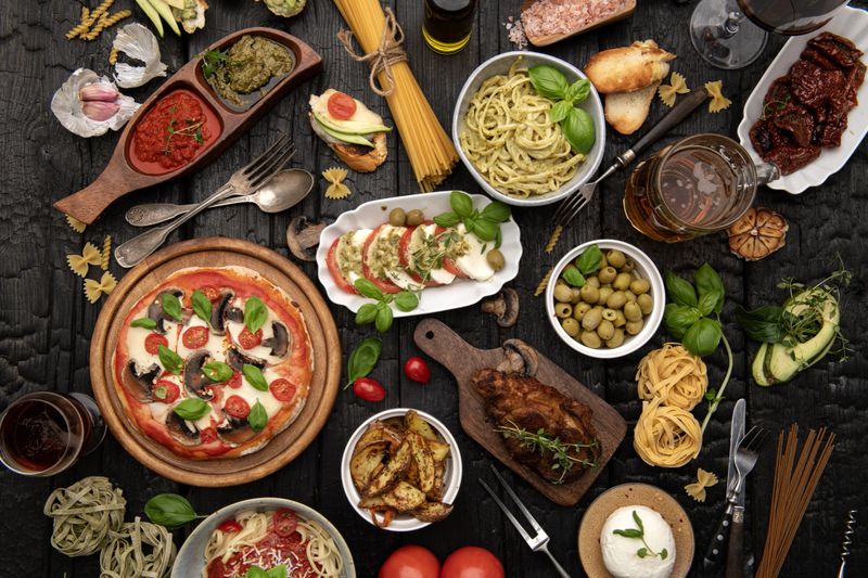 Top view of homemade Italian meal with pizza, pasta with pesto, grilled meat, olives, roasted potatoes and wine on dark rustic background. Warm Mediterranean atmosphere.