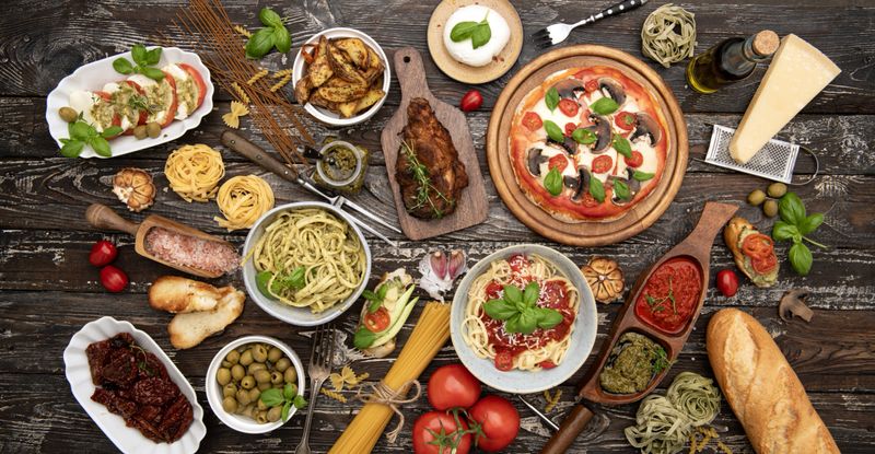 Top view of Italian food composition with pizza, pasta with pesto, caprese salad, grilled meat, olives, and parmesan cheese on rustic wooden table.Keywords (25):