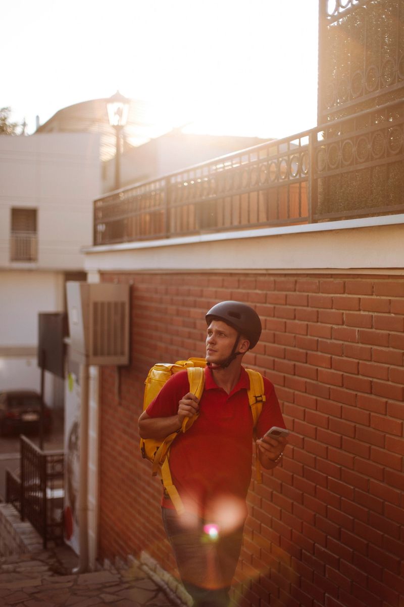 A delivery courier wearing a red polo and helmet, clutching a smartphone while walking along a brick alley at sunset; Warm light and lens flare highlight a focused, urban day-in-the-life moment
