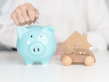 Hand putting coin in piggy bank with wooden car and house model.