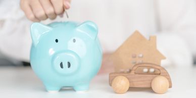 A hand placing a coin into a blue piggy bank next to wooden house and car models.