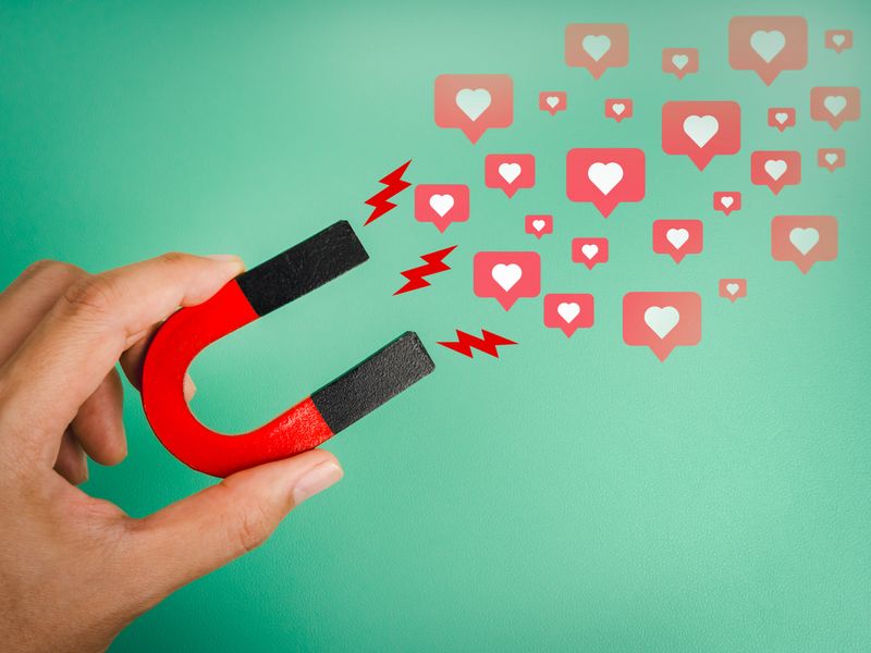 Hand holding red magnet attracting social media like icons, symbolizing digital marketing, content engagement, social influence, and online audience connection on green background.