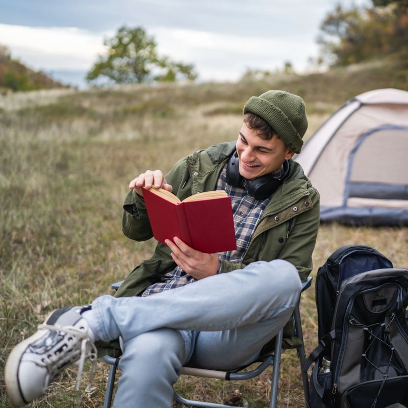 Young man wearing a beanie and headphones relaxing in a folding chair, reading a book intently, next to a camping tent and backpack in an outdoor wilderness setting