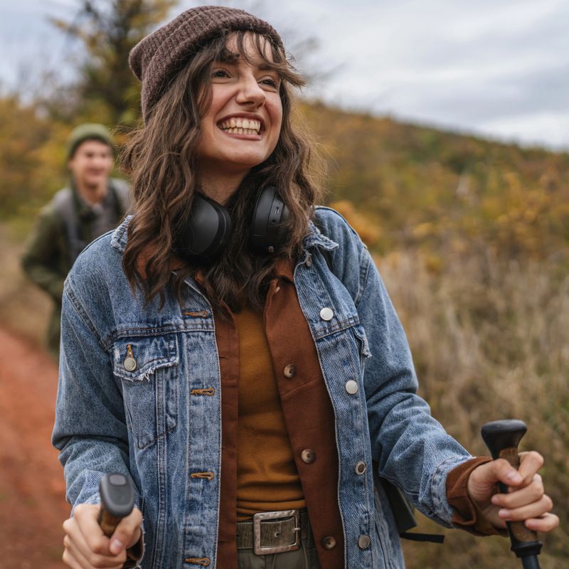 Happy young woman smiling warmly and looking ahead, enjoying nature while hiking along a rural autumn trail with a partner, headphones around her neck, holding trekking poles