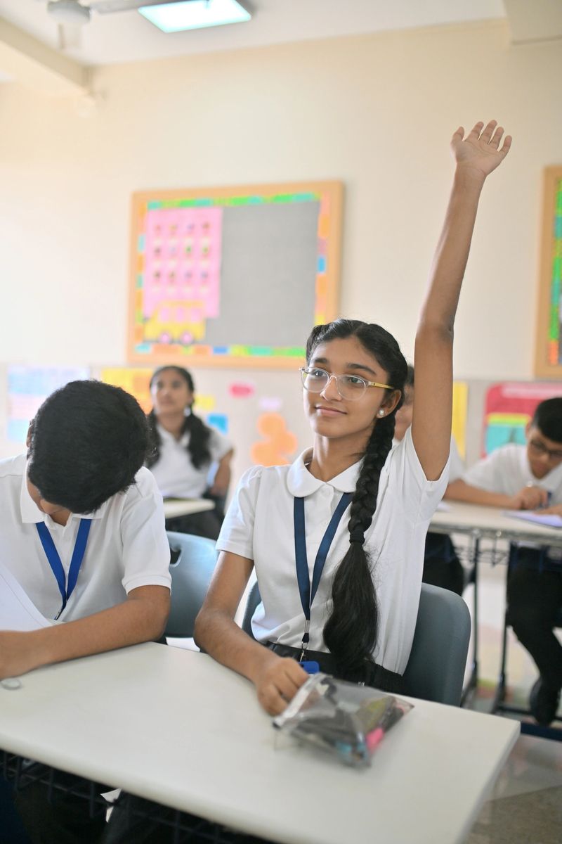 Confident Indian student in school uniform sitting at her desk, eagerly raising her hand to answer the teacher during class. Other classmates are focused on their work in the background. Concept of active participation, learning attitude, girl education, and classroom engagement in an Indian school.