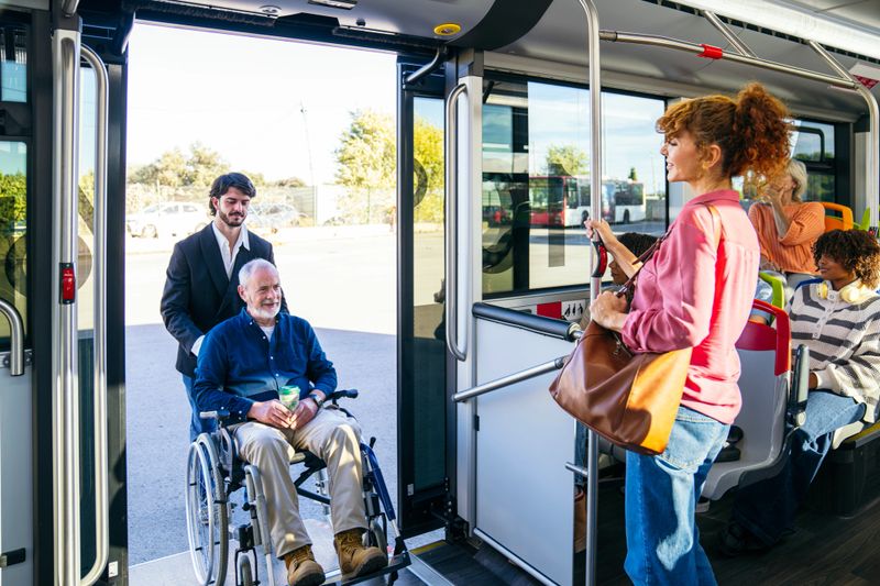 Young man assisting a smiling senior adult in a wheelchair boarding a public city bus, showing accessibility and care
